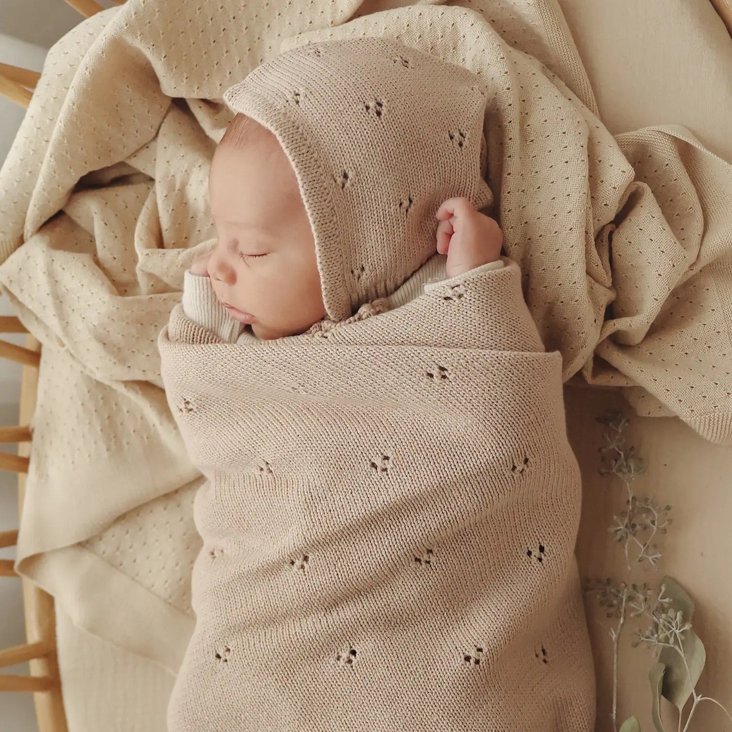 Newborn baby wrapped in a beige knitted swaddle with matching bonnet, surrounded by soft textures.