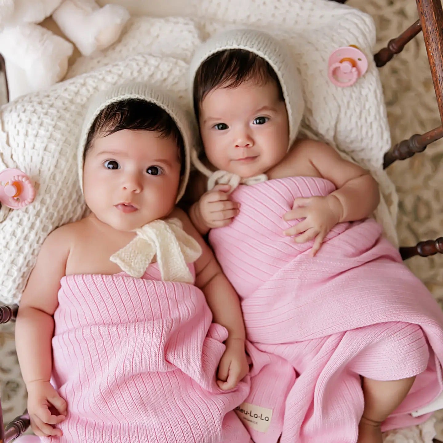 Two babies wrapped in pink blankets with white bonnets on a wooden crib.