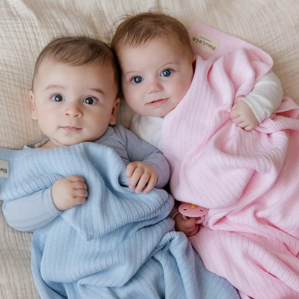 Two babies wrapped in blue and pink blankets on a light-colored surface.