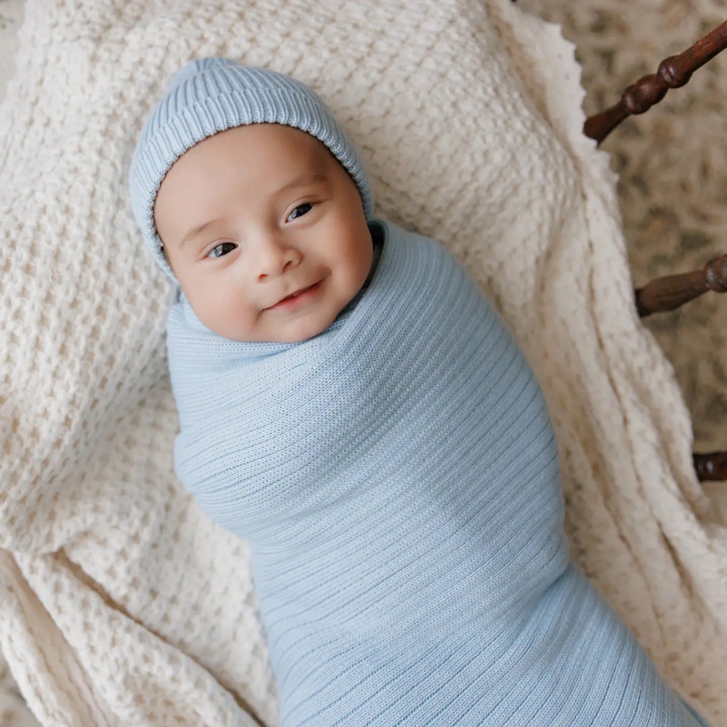 Baby wrapped in a light blue swaddle and hat, lying on a textured beige blanket.